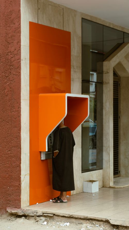 Free A person in casual attire uses an ATM on a street corner with an orange accent wall. Stock Photo