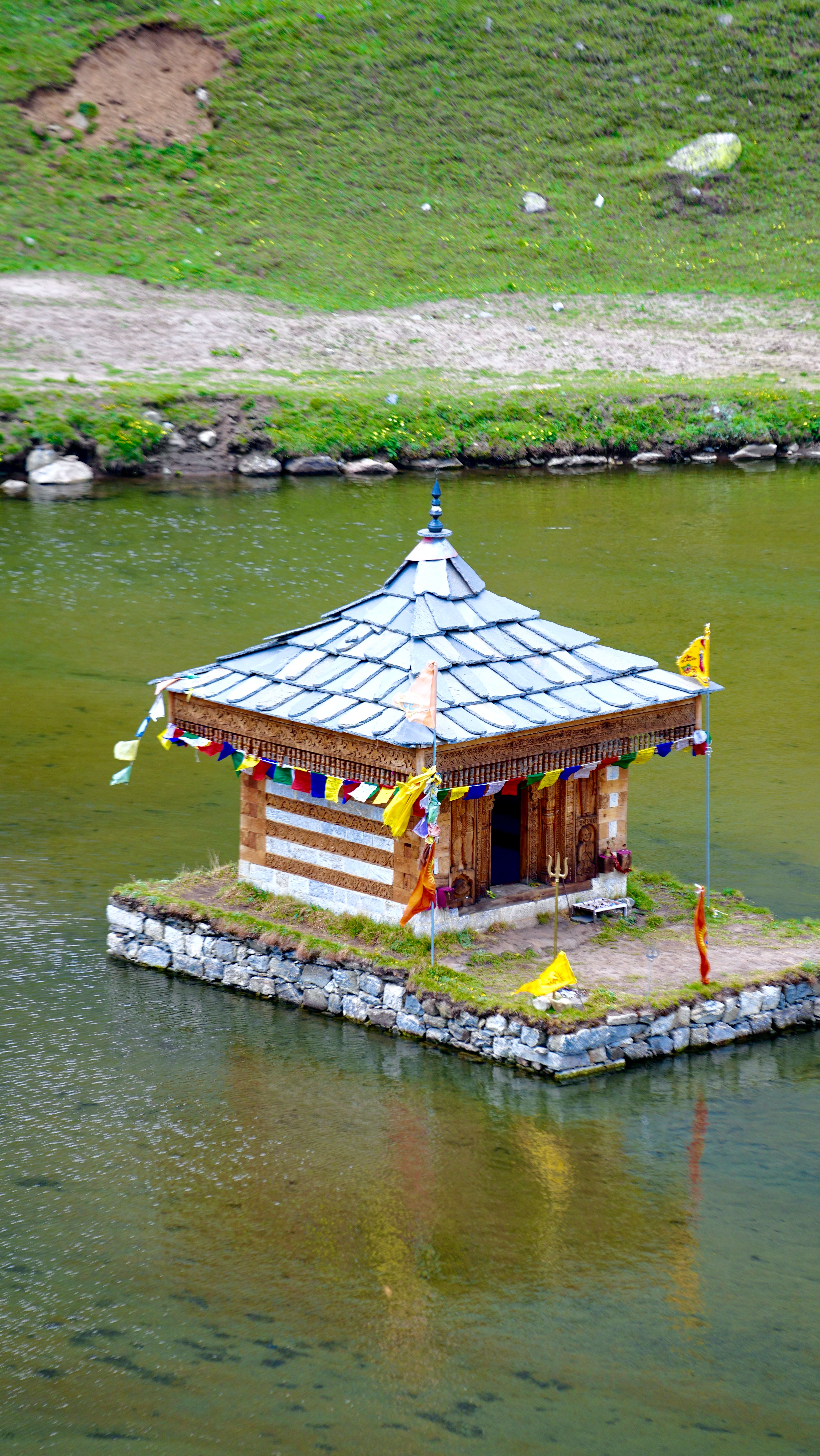 A traditional wooden hut sits on a serene lake, surrounded by lush greenery, under a summer sky.