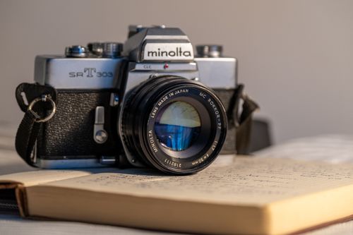 Free A classic Minolta SR-T 303 analog camera with leather strap on a bed alongside an open notebook, evoking nostalgia. Stock Photo