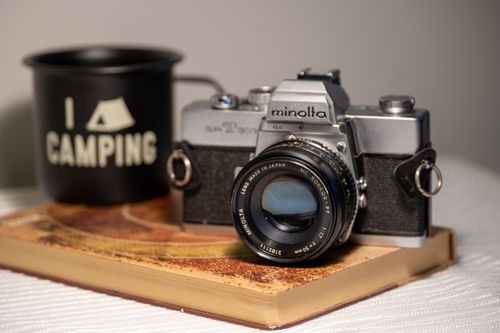 Free Retro Minolta film camera on book with a camping-themed mug, showcasing nostalgia. Stock Photo
