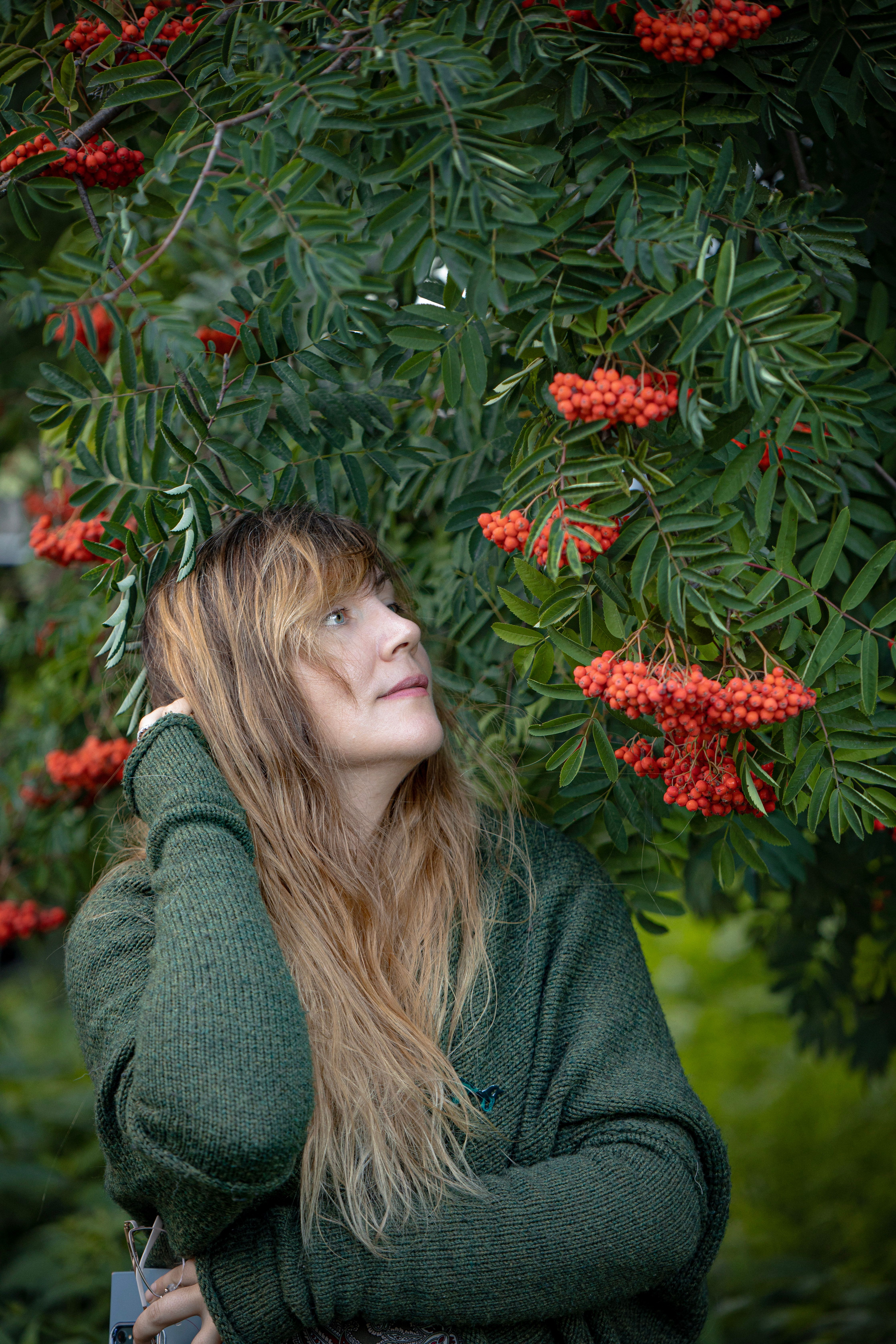 A woman in a green sweater gazes up at vibrant rowan berries hanging from green foliage.