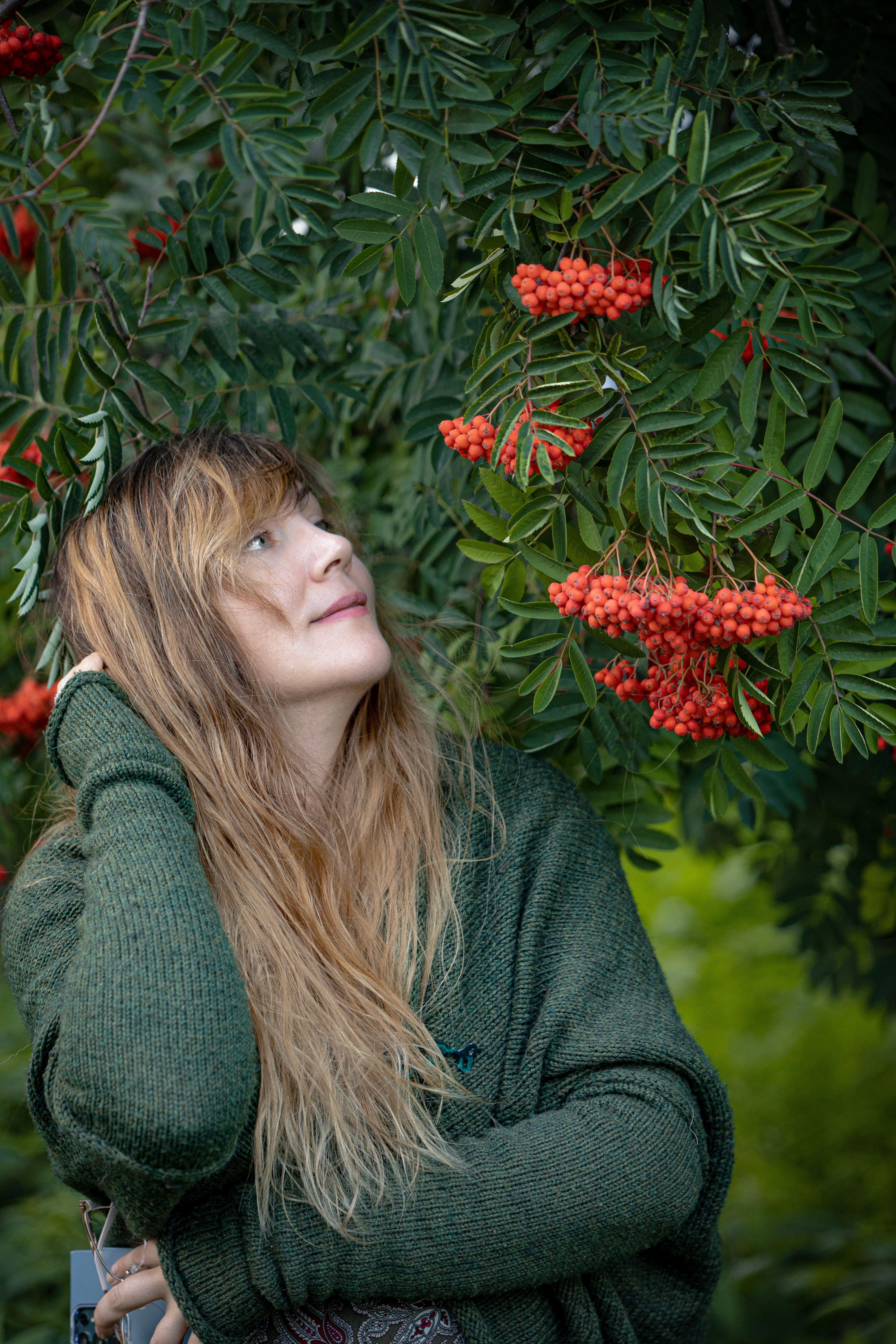 A woman in a green sweater looks up at vibrant rowan berries on a tree branch outdoors.