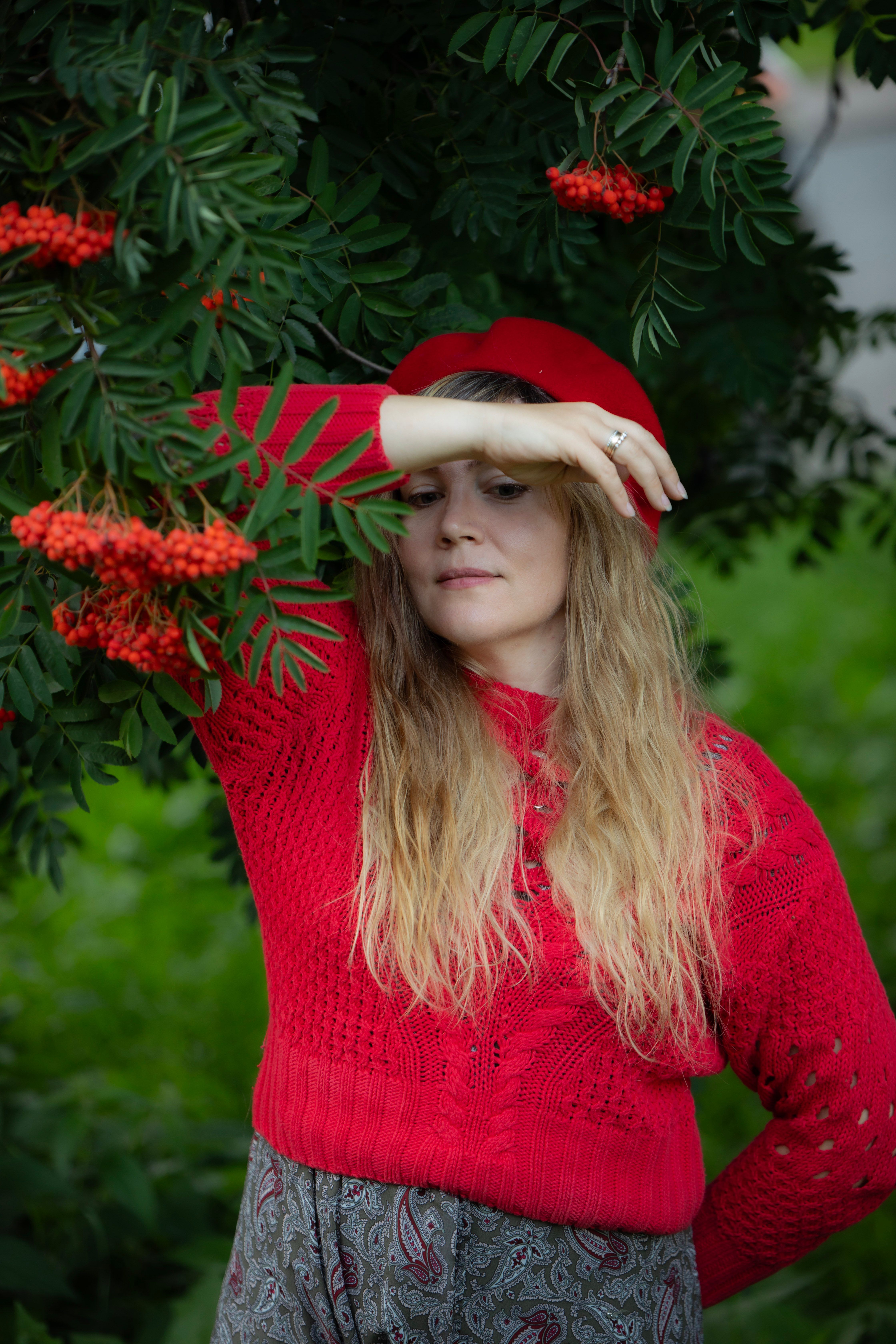 A woman in a red sweater stands gracefully beside rowan trees in a serene outdoor setting.