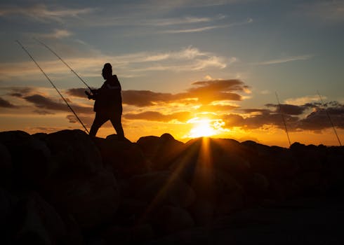 A fisherman silhouetted against a vibrant sunset over the ocean, casting a serene scene.