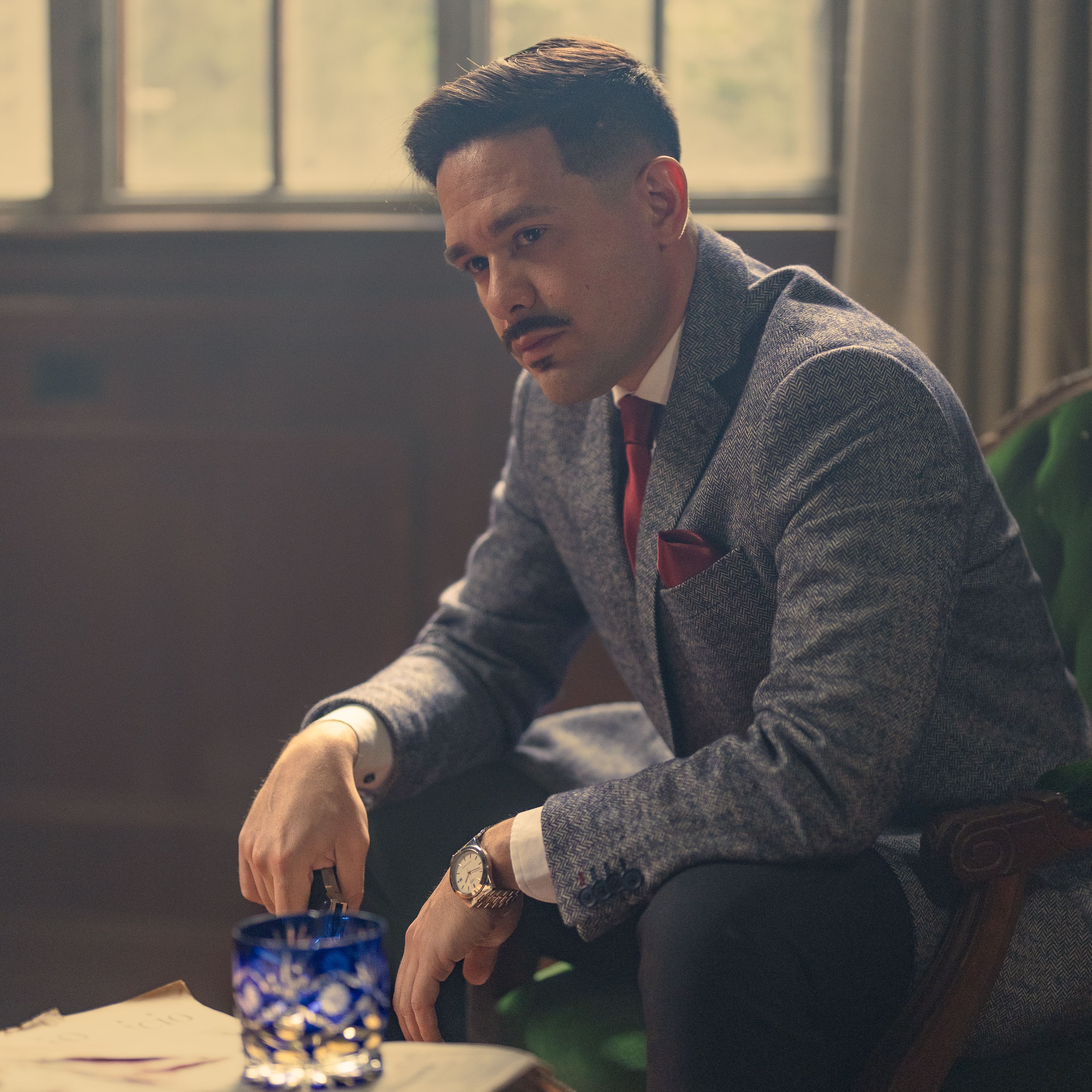 Elegant man in formal attire sits pensively by a window, exuding sophistication.