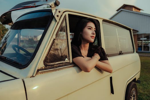 A woman leaning out of a vintage car window, enjoying the sunset outdoors.