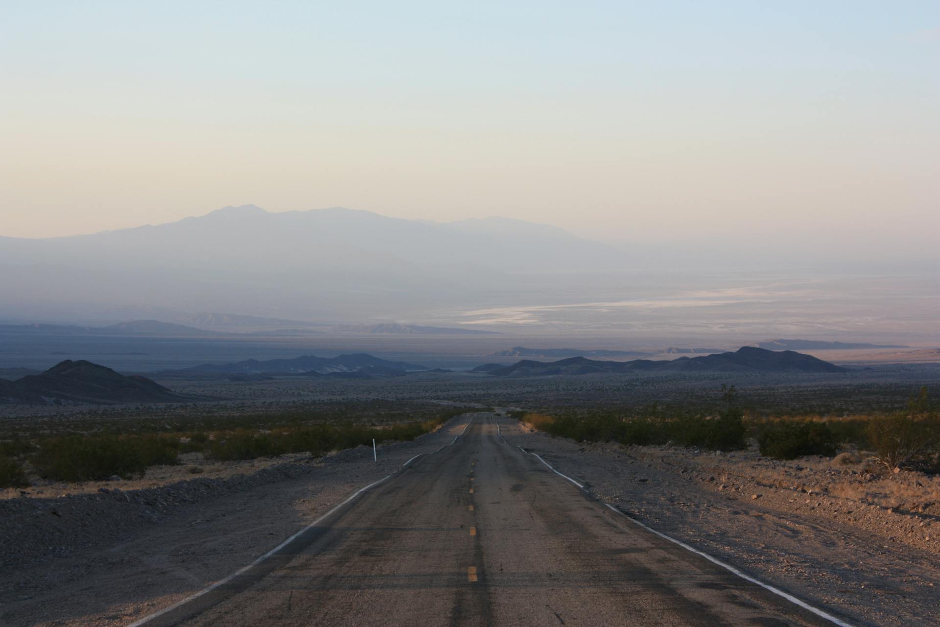 Long desert road leading to distant mountains at sunrise, with a serene and remote landscape.