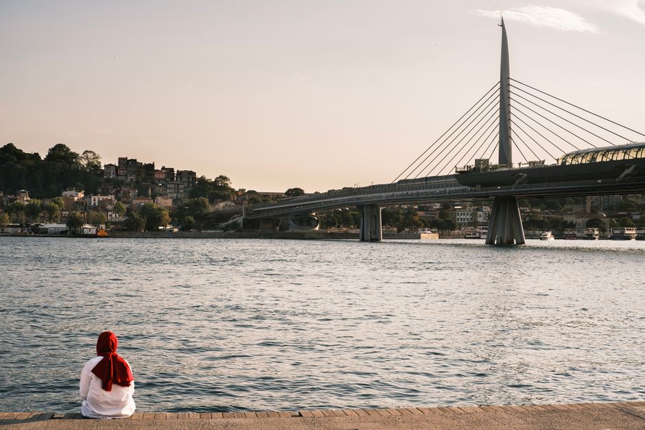 Person sits by the Golden Horn with Haliç Metro Bridge at sunset in Istanbul, Türkiye.