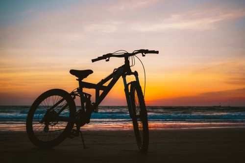 Free Silhouetted bicycle against a vibrant sunset over a calm beach, perfect for leisure and travel themes. Stock Photo