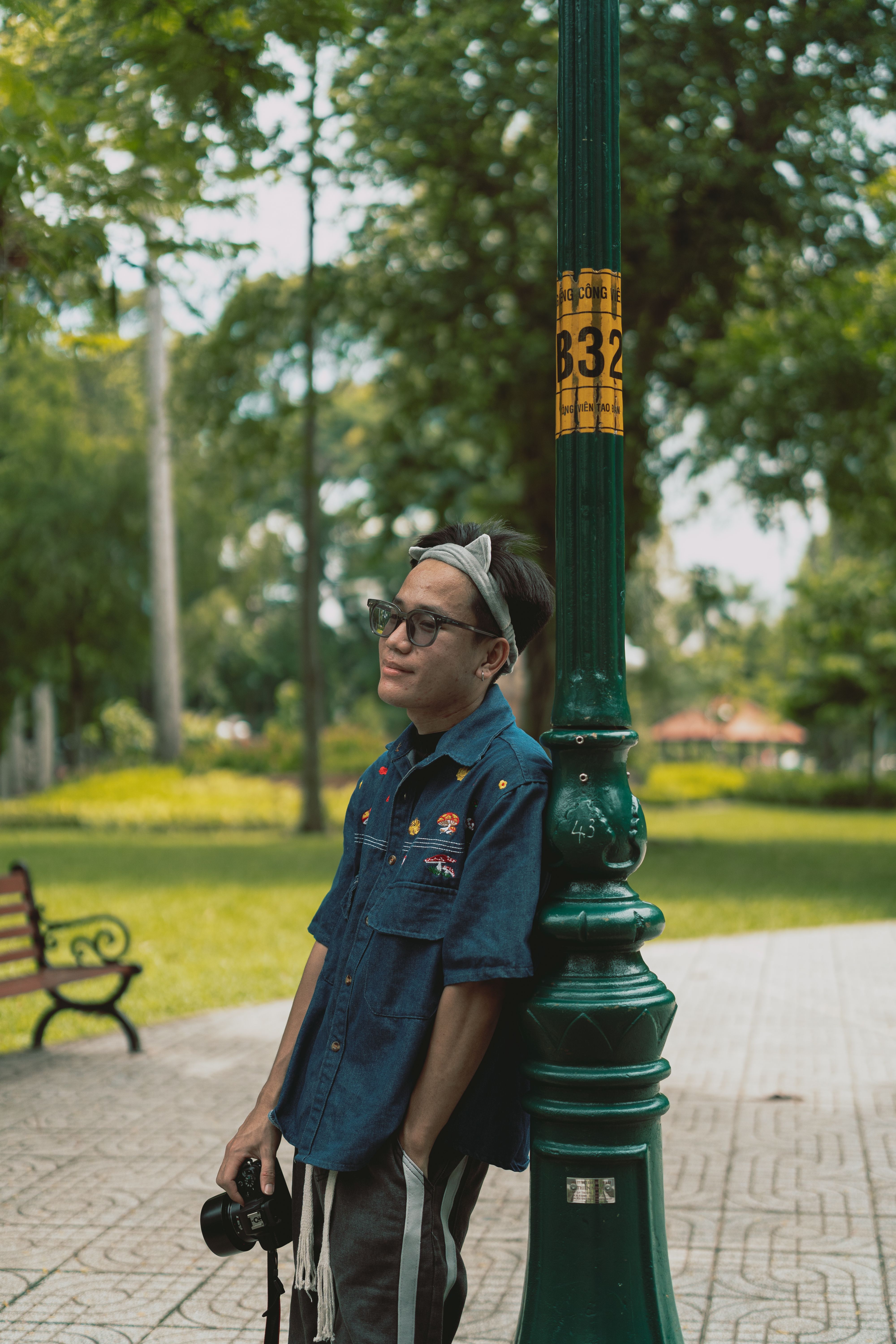 A relaxed man stands by a lamppost in a lush urban park, holding a camera.
