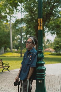 A relaxed man stands by a lamppost in a lush urban park, holding a camera.