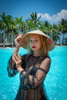 A woman in a sheer dress and Asian hat poses by a tropical pool surrounded by palm trees.