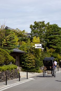 A classic black rickshaw travels through a traditional Japanese village surrounded by lush greenery and architecture.