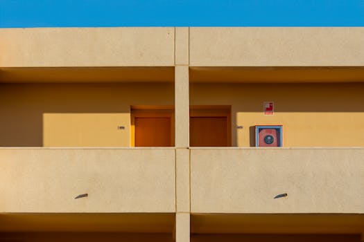 Modern minimalist apartment facade featuring two brown doors and a fire hose reel. Clean lines and bright colors.