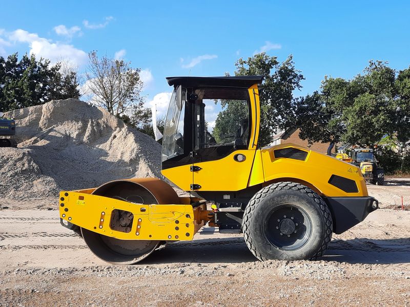 Close-up of a yellow road roller on a construction site with gravel and trees in the background