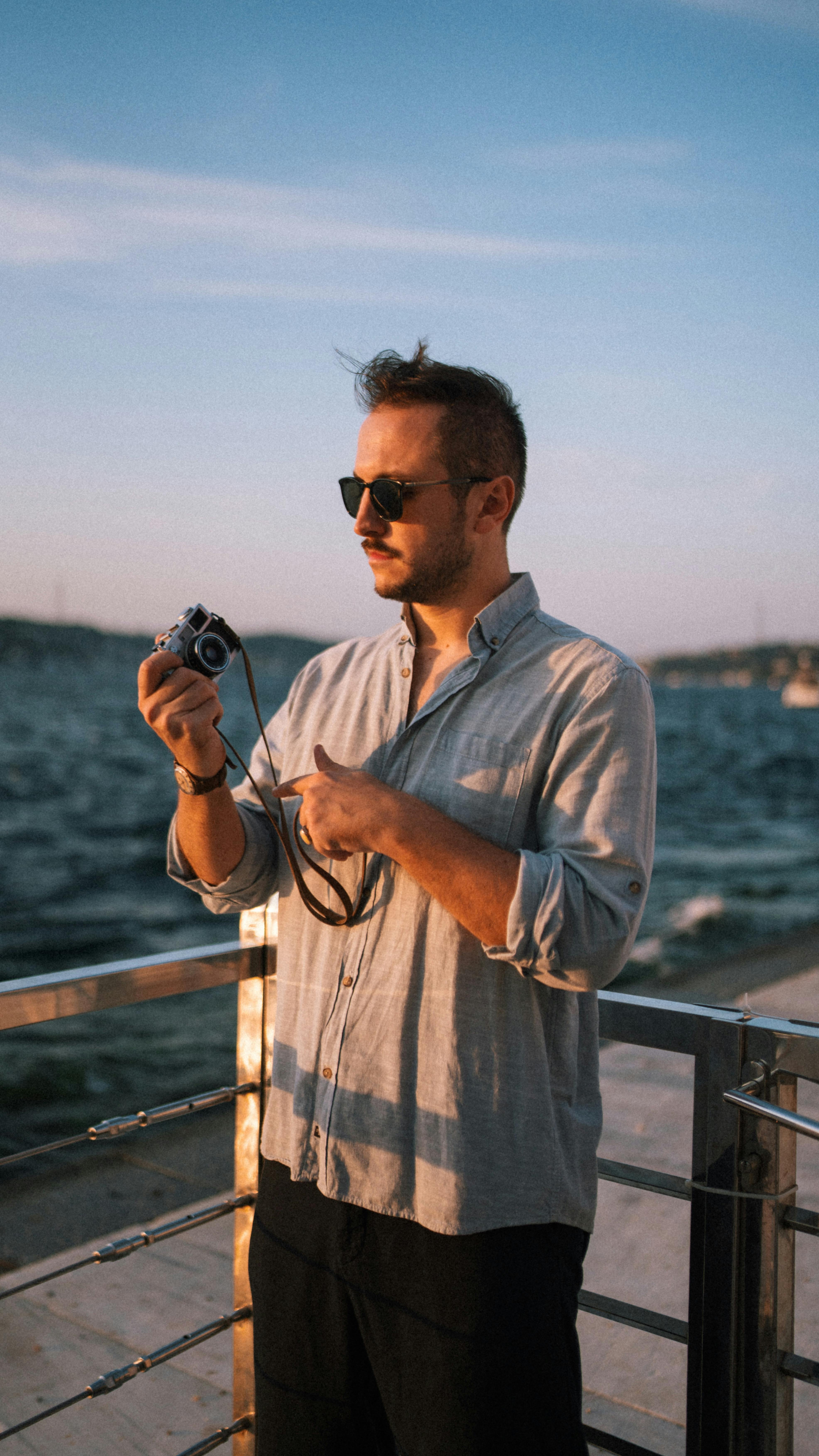 Man with sunglasses holding a camera by the waterfront at sunset, embracing leisure.