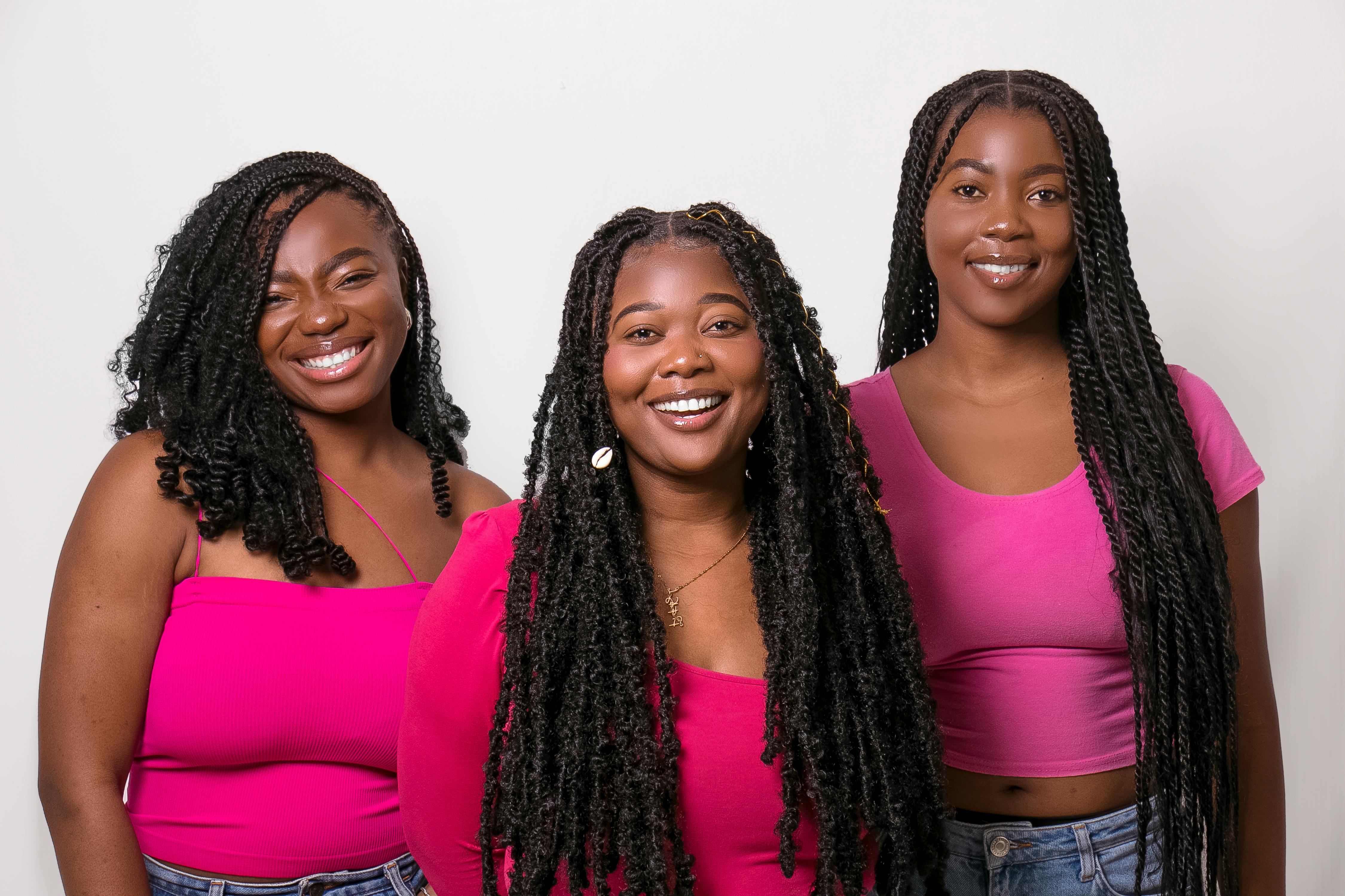 Three young women with braided hairstyles smiling confidently in pink attire.