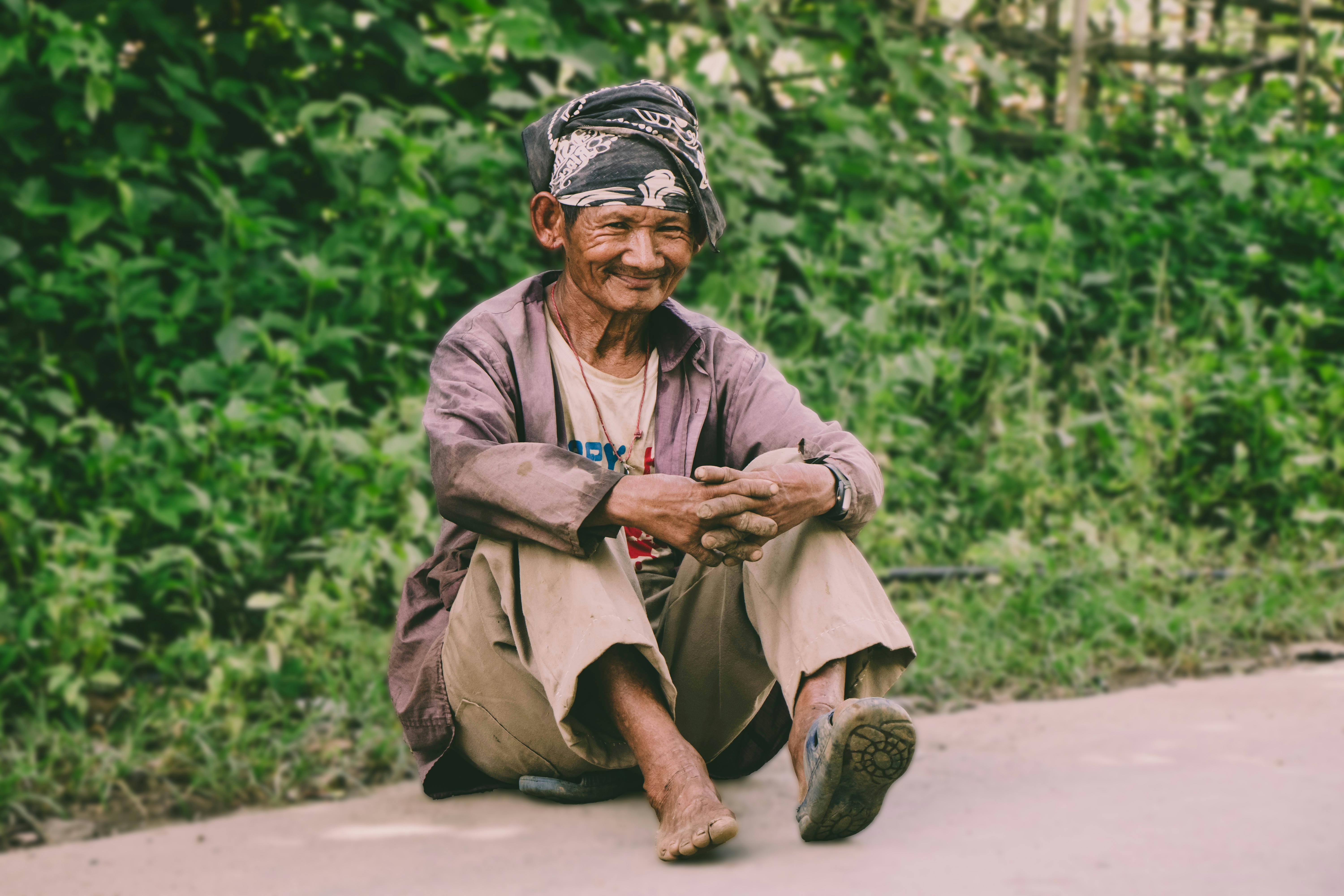 一位老人赤脚坐在绿树成荫的小路上，脸上洋溢着温暖的笑容