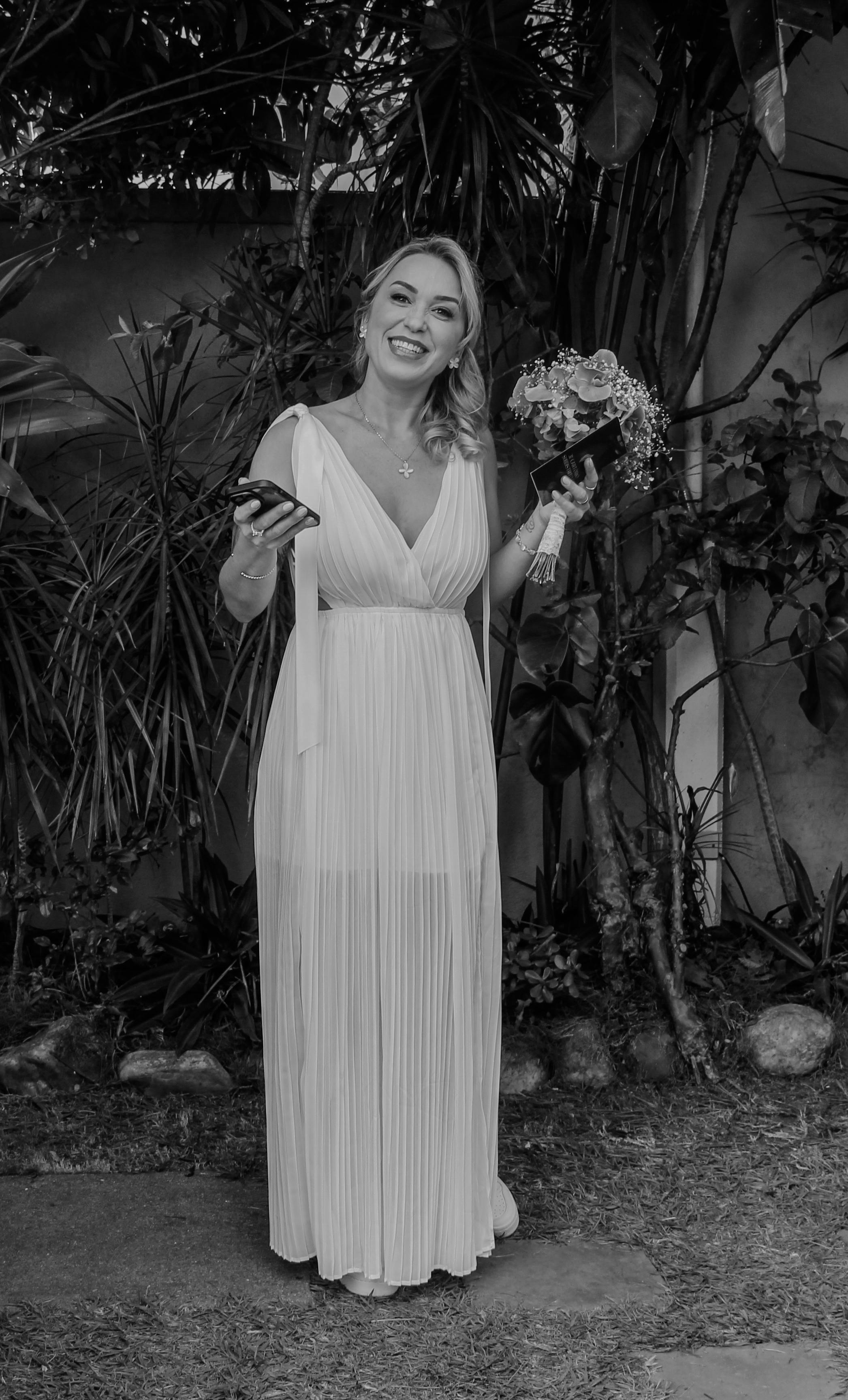 Black and white photo of a smiling bride holding a bouquet and phone in a garden.