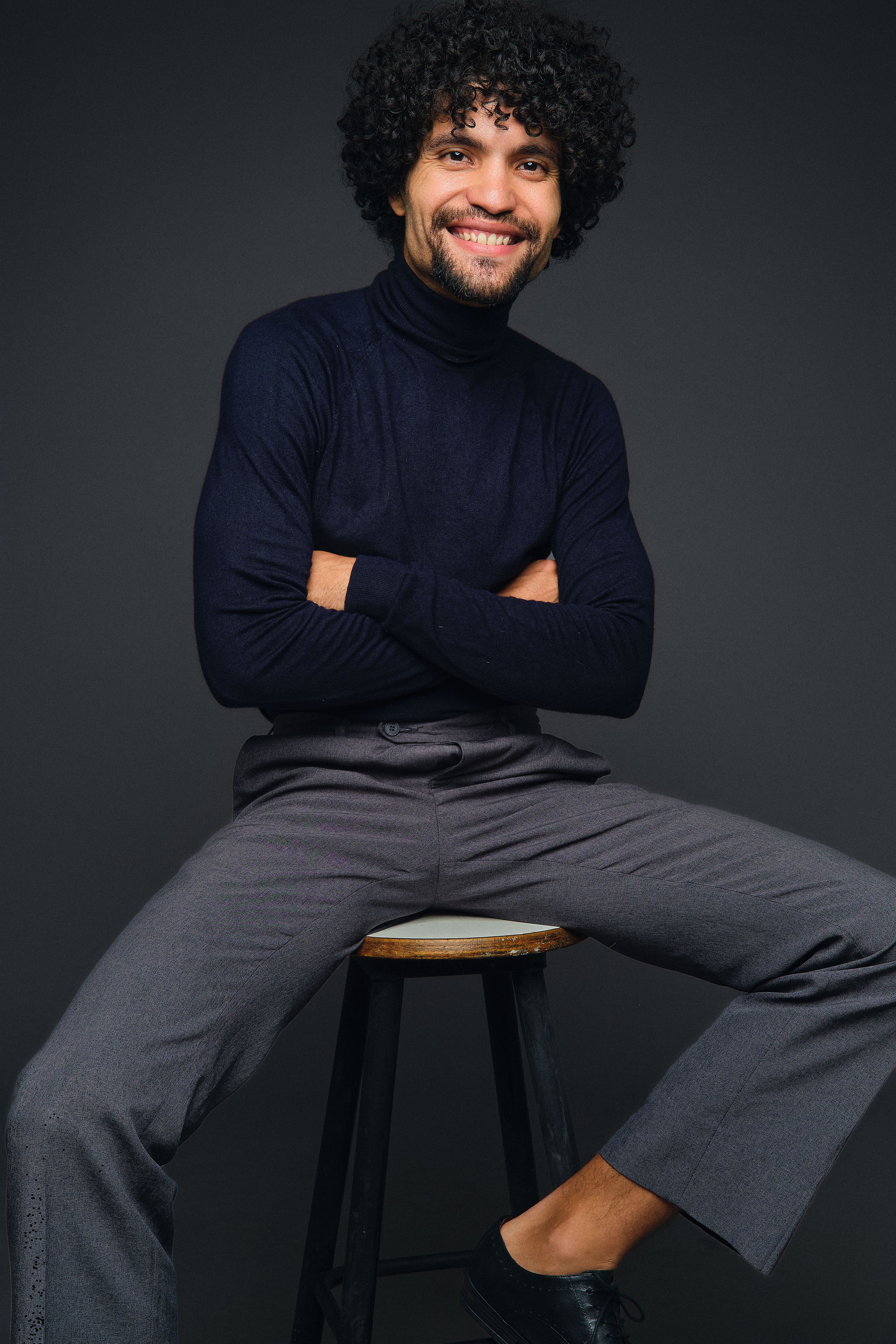 Portrait of a stylish man sitting on a stool with a confident smile in a studio setting.