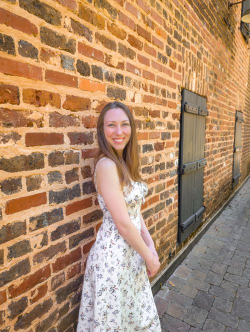 Free Young woman wearing a floral dress smiles brightly against a rustic brick wall in an urban setting. Stock Photo