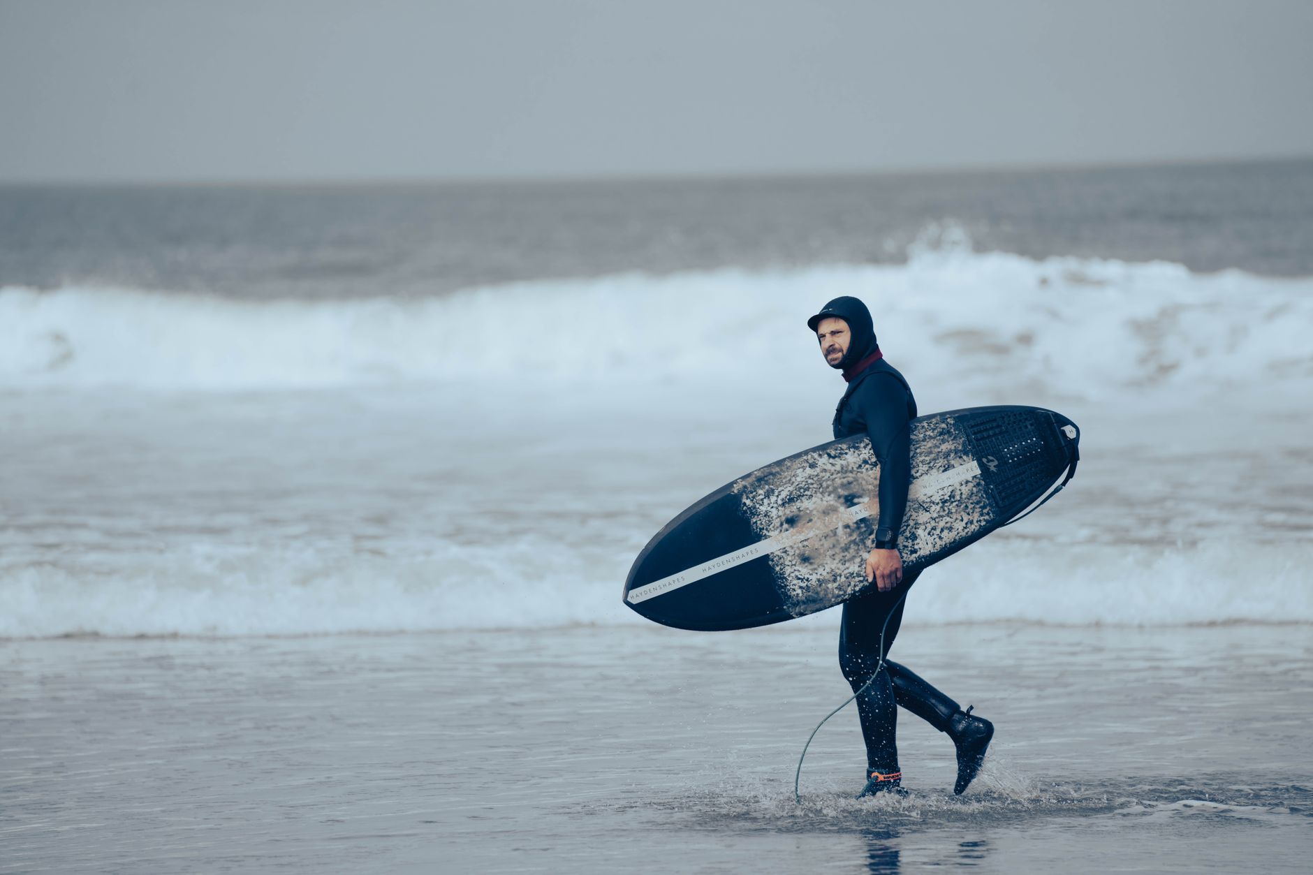 Man in wetsuit with surfboard by ocean, ready for surfing adventure.