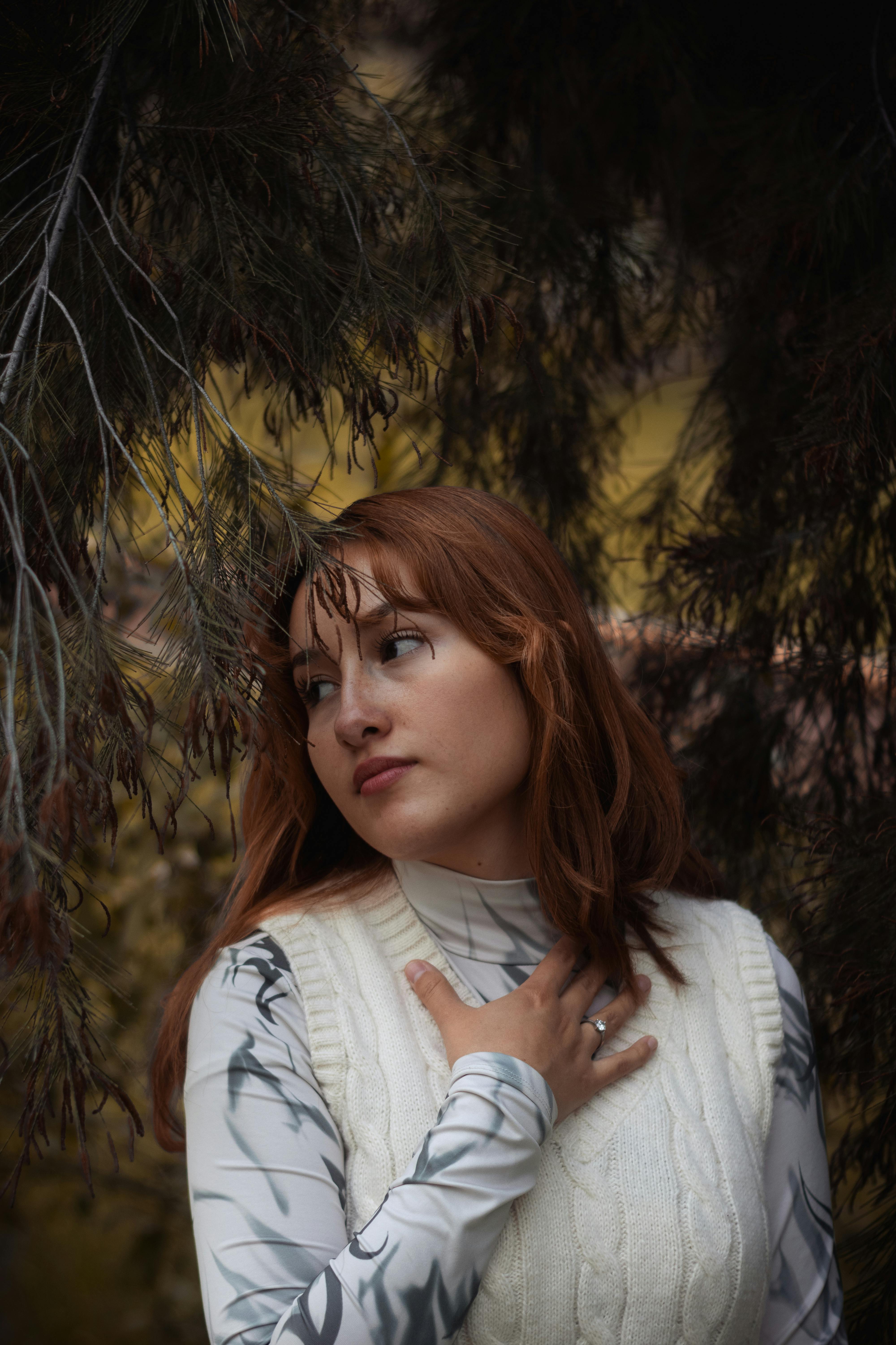 A thoughtful woman with auburn hair surrounded by pine branches in a fall setting.
