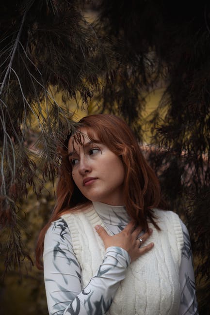 A thoughtful woman with auburn hair surrounded by pine branches in a fall setting.