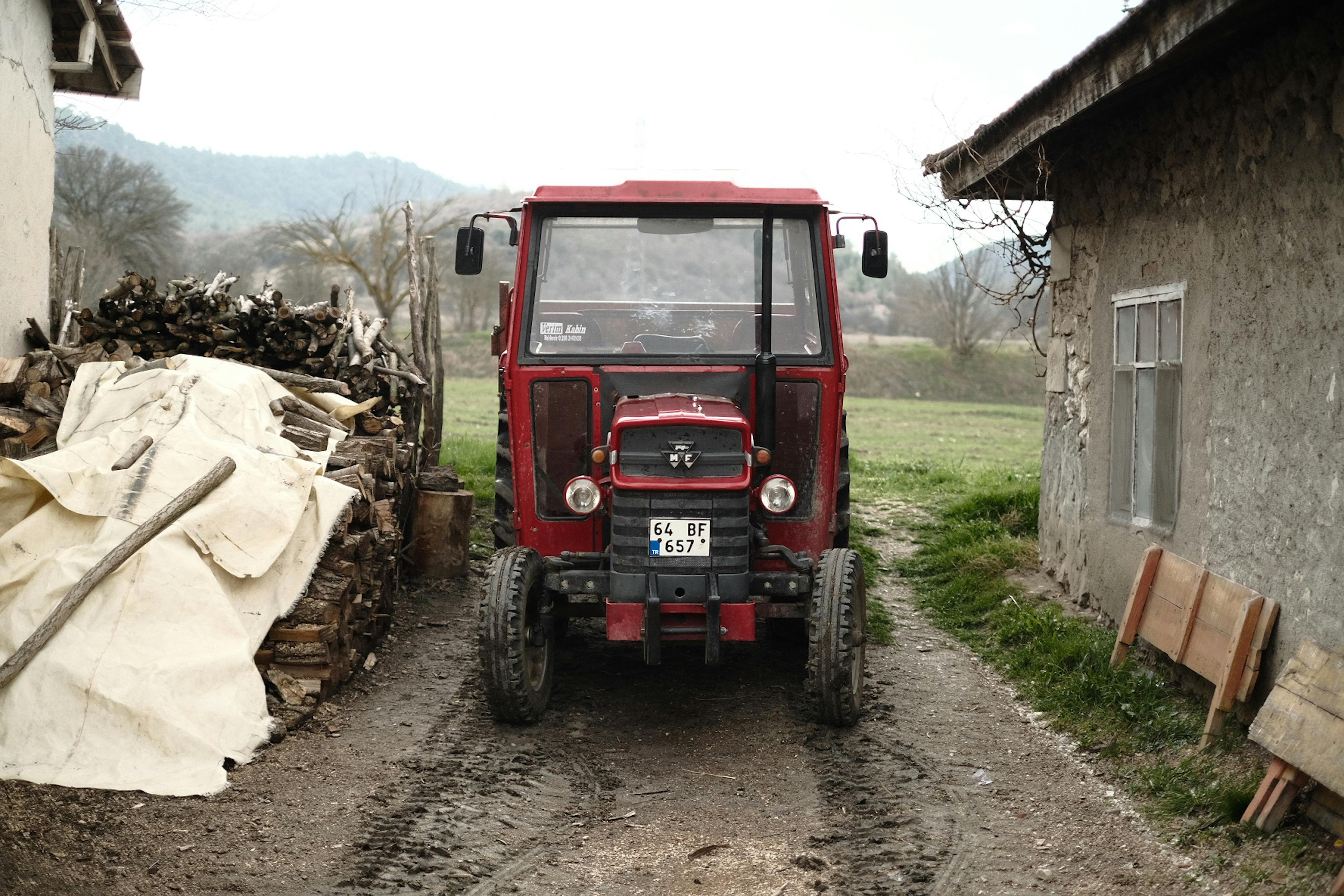 A red tractor parked on a rural farm road beside wooden logs and a stone house.
