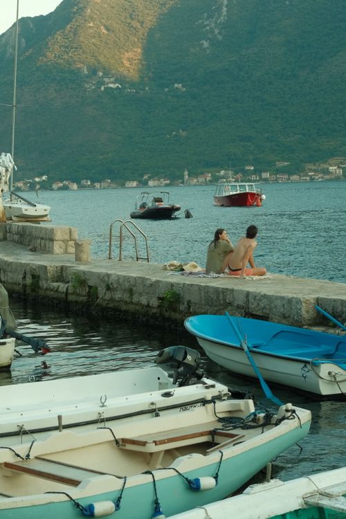Free Two people relaxing by the harbor with boats and a mountainous background on a sunny day. Stock Photo