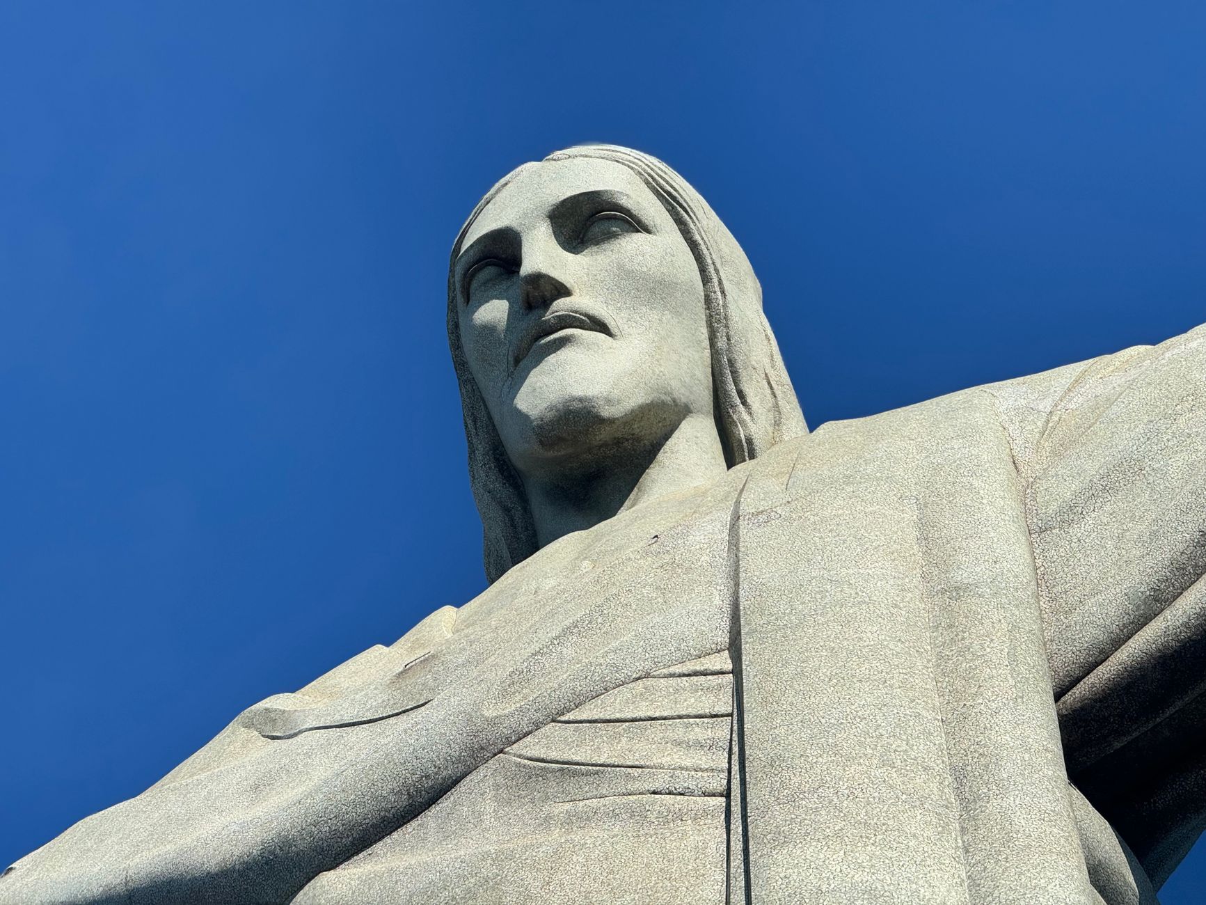 A low-angle close-up of the face and outstretched arm of the Christ the Redeemer statue against a deep blue sky on Corcovado