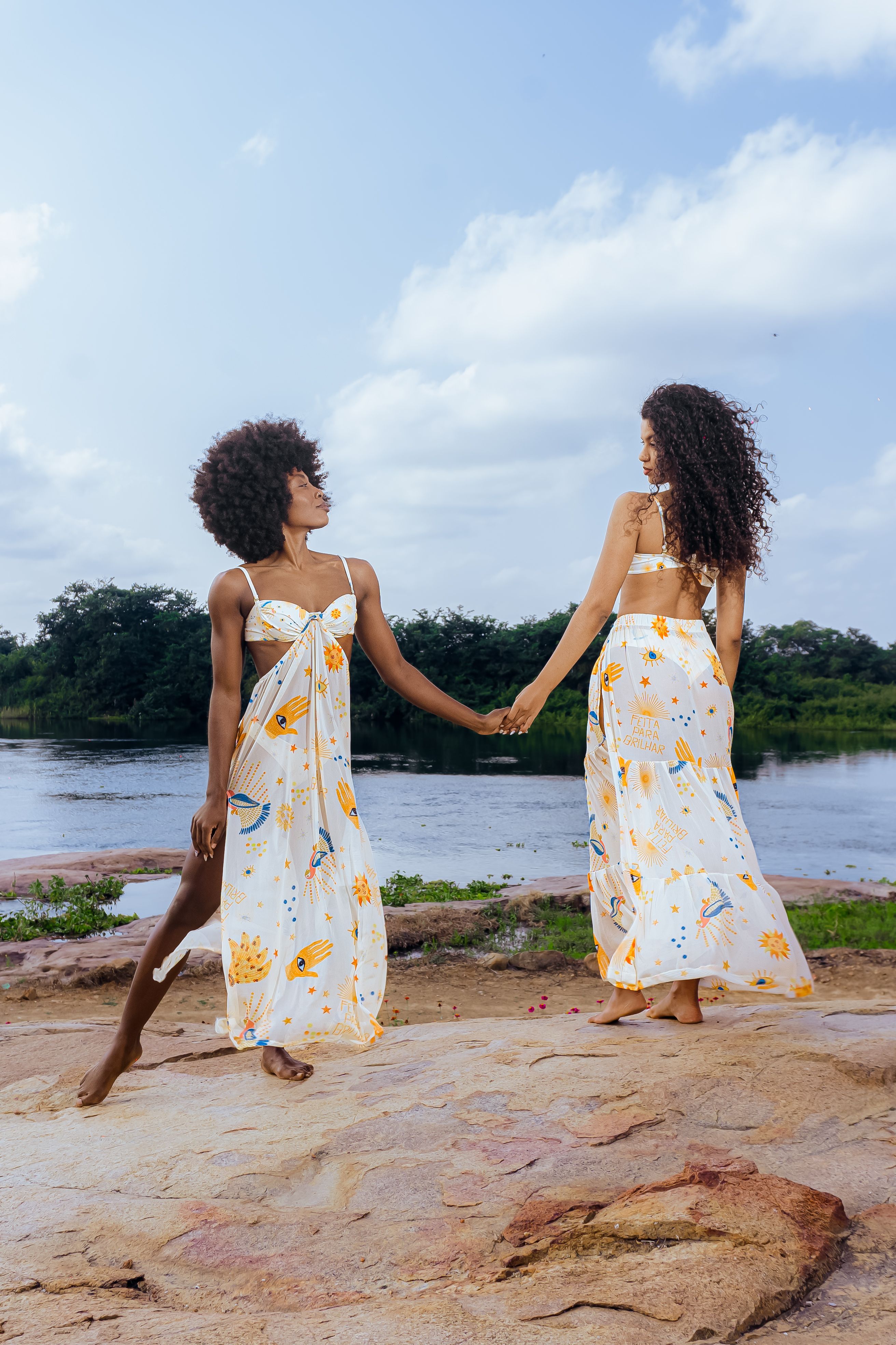 Two women holding hands, wearing summer dresses by a riverbank, enjoying a sunny day.