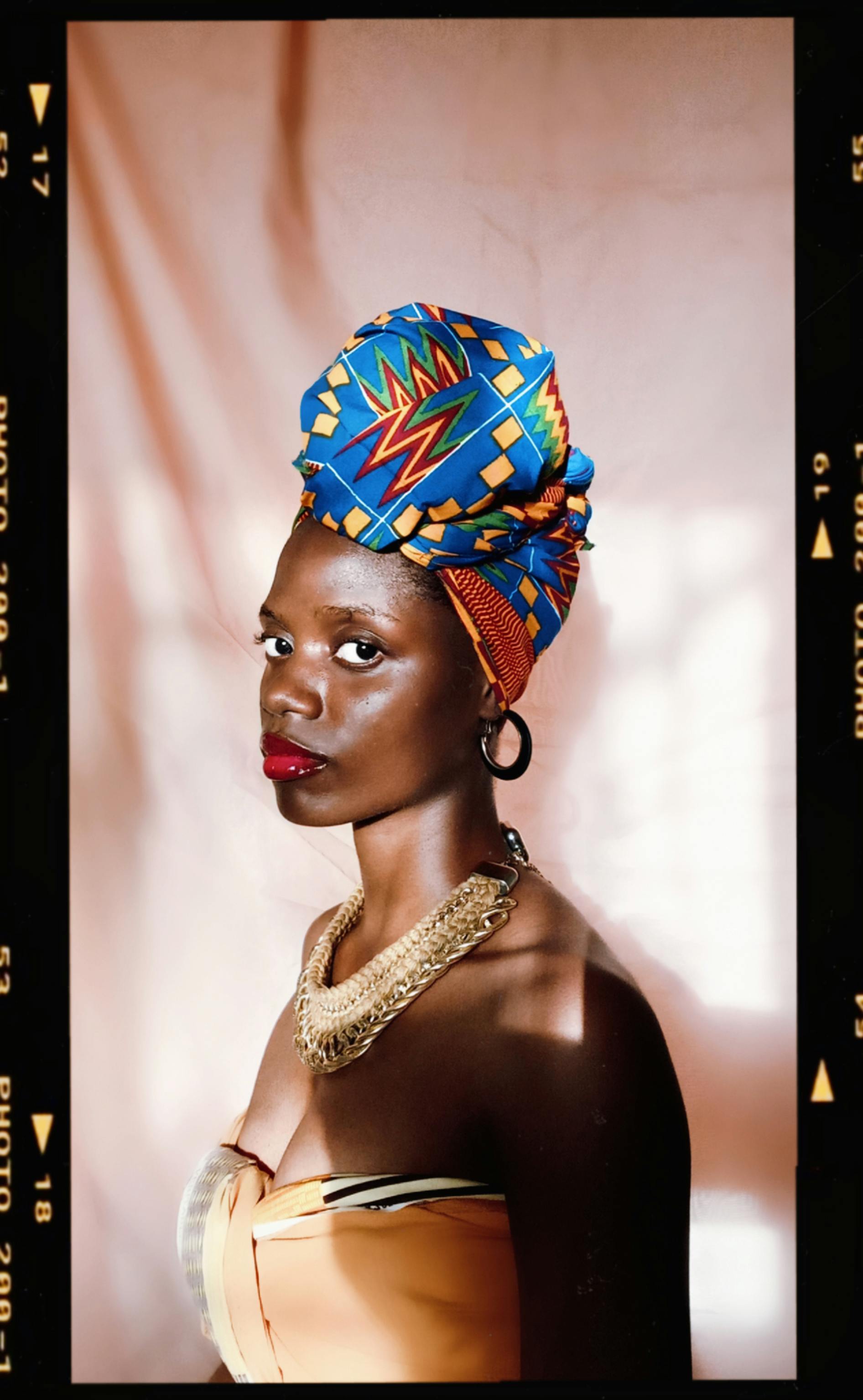 Portrait of a woman wearing a vibrant African headwrap and jewelry against a soft backdrop.