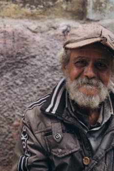 A warm portrait of an elderly man with a beard and cap, sitting outdoors in Alexandria, Egypt.