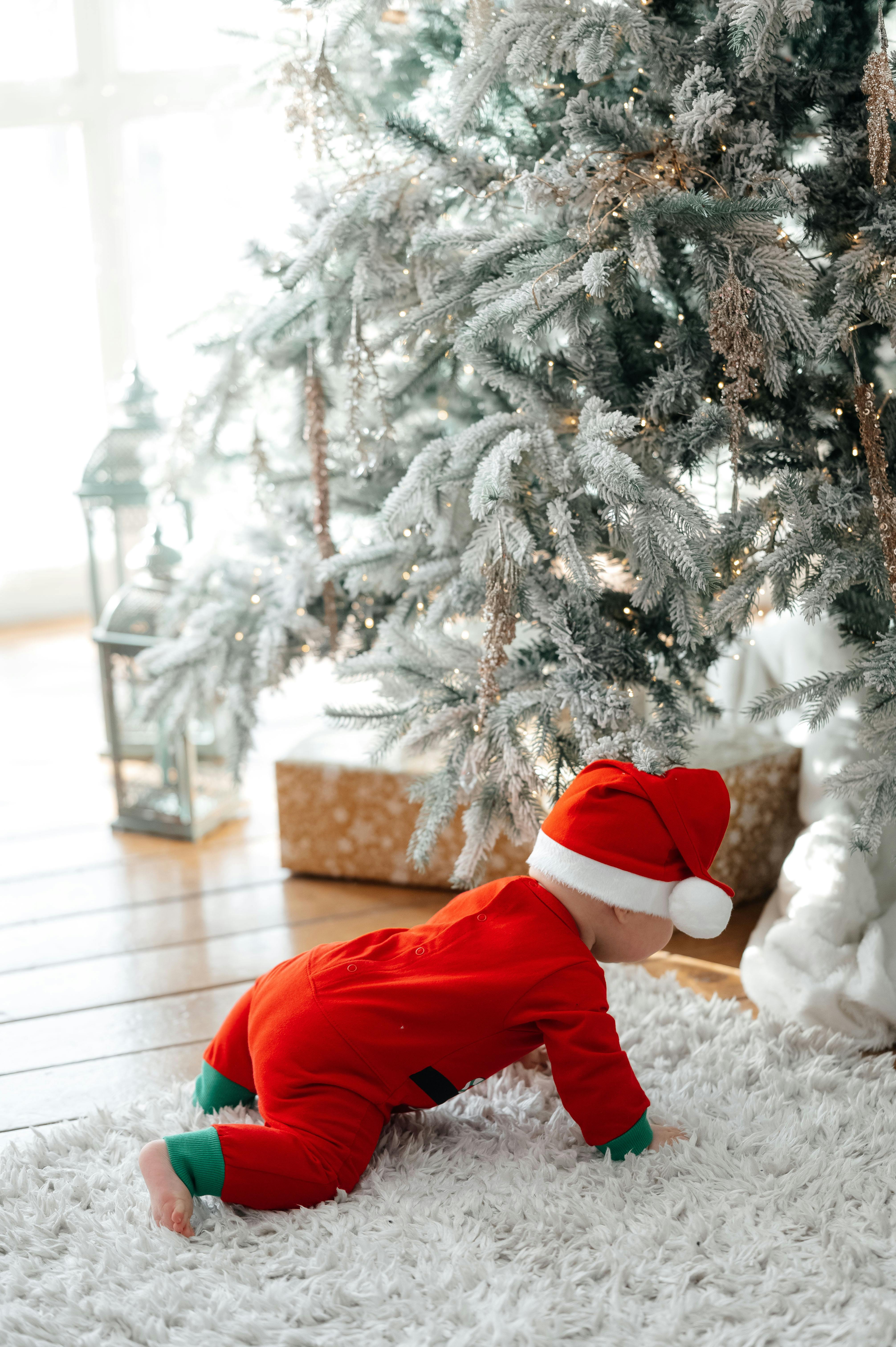 A cute baby in a Santa suit crawls near a frosted Christmas tree indoors.