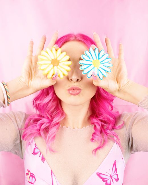 Portrait of a woman with vibrant pink hair holding daisy-shaped cookies. Bright and playful style.