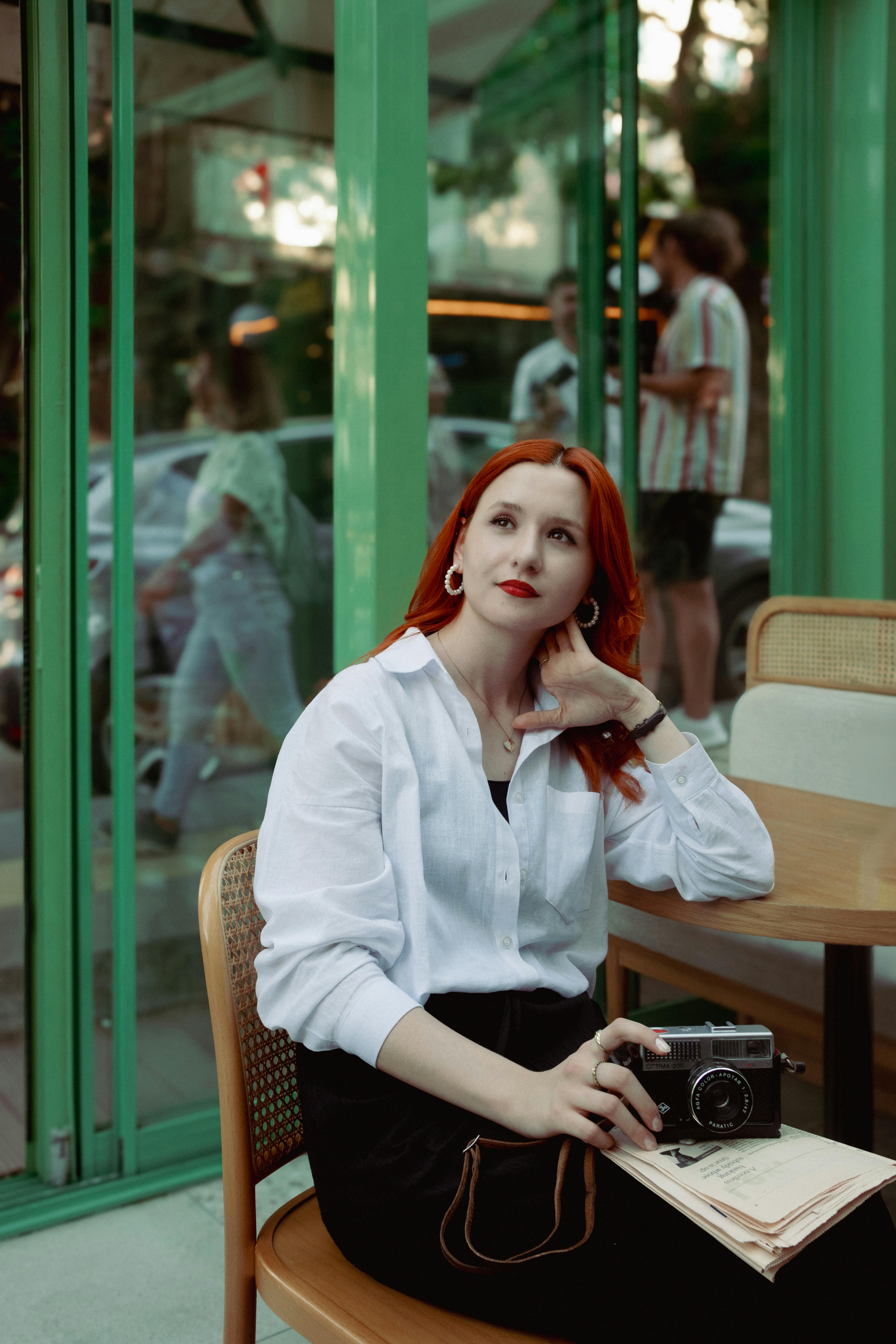 Elegant redhead woman sitting in a café with a vintage camera. Stylish and thoughtful.