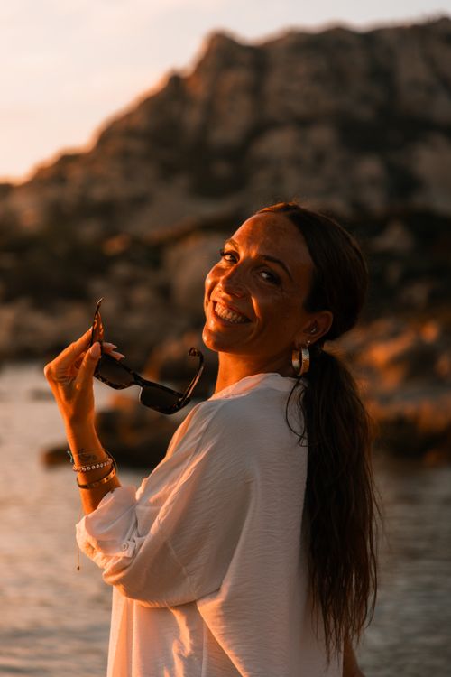 Free A woman enjoys the golden hour at a beach with rocky formations, holding sunglasses and smiling. Stock Photo