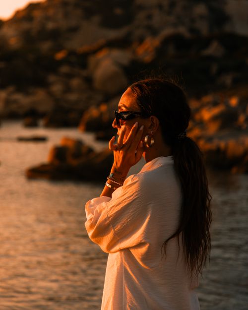 Free Serene woman in sunglasses savors sunset at rocky beach with warm tones. Stock Photo