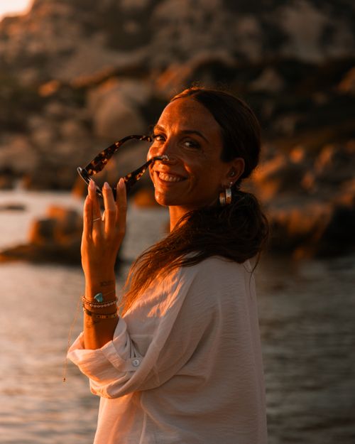 Free A smiling woman holding sunglasses by the beach during a warm sunset. Stock Photo