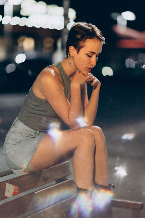 Free A young woman sitting thoughtfully in an urban setting at night with artistic bokeh lights. Stock Photo
