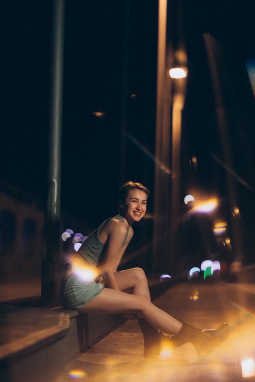 Free A woman smiling while seated on a city street at night, surrounded by urban lights. Stock Photo