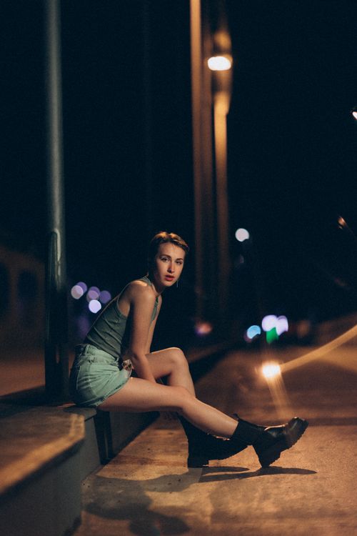 Free A fashionable woman sits on a city street at night, creating a moody urban scene. Stock Photo