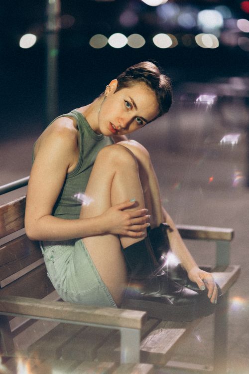 Free Elegant woman sitting on a bench in casual attire at night with soft bokeh lights. Stock Photo