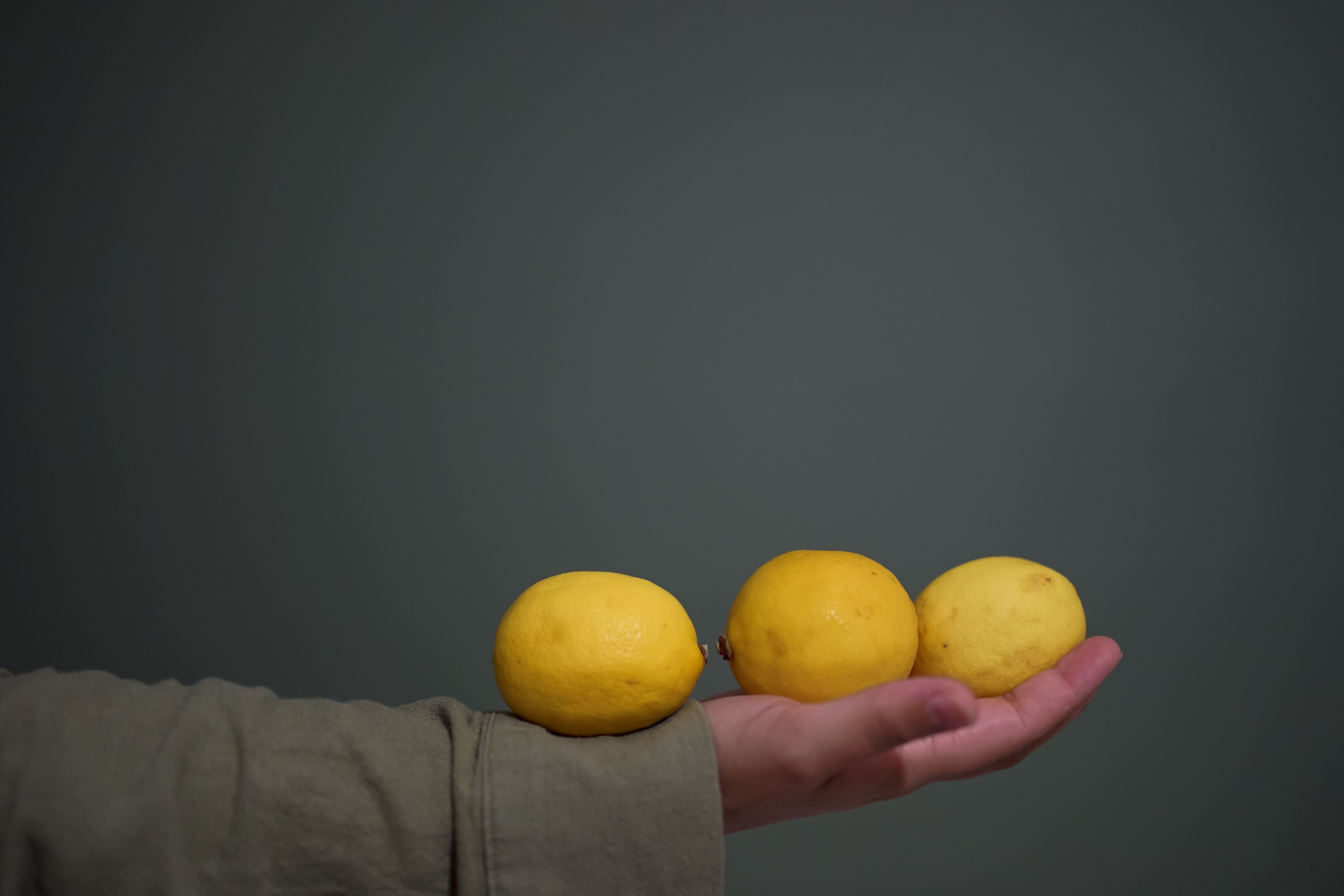 A hand holding three ripe yellow lemons against a minimalist backdrop.