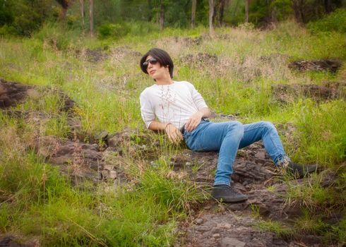 A young man in casual attire relaxing on a grassy, rocky outdoor setting.