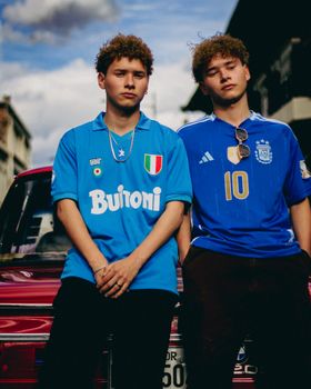 Two young men wearing football jerseys pose confidently beside a car outdoors.
