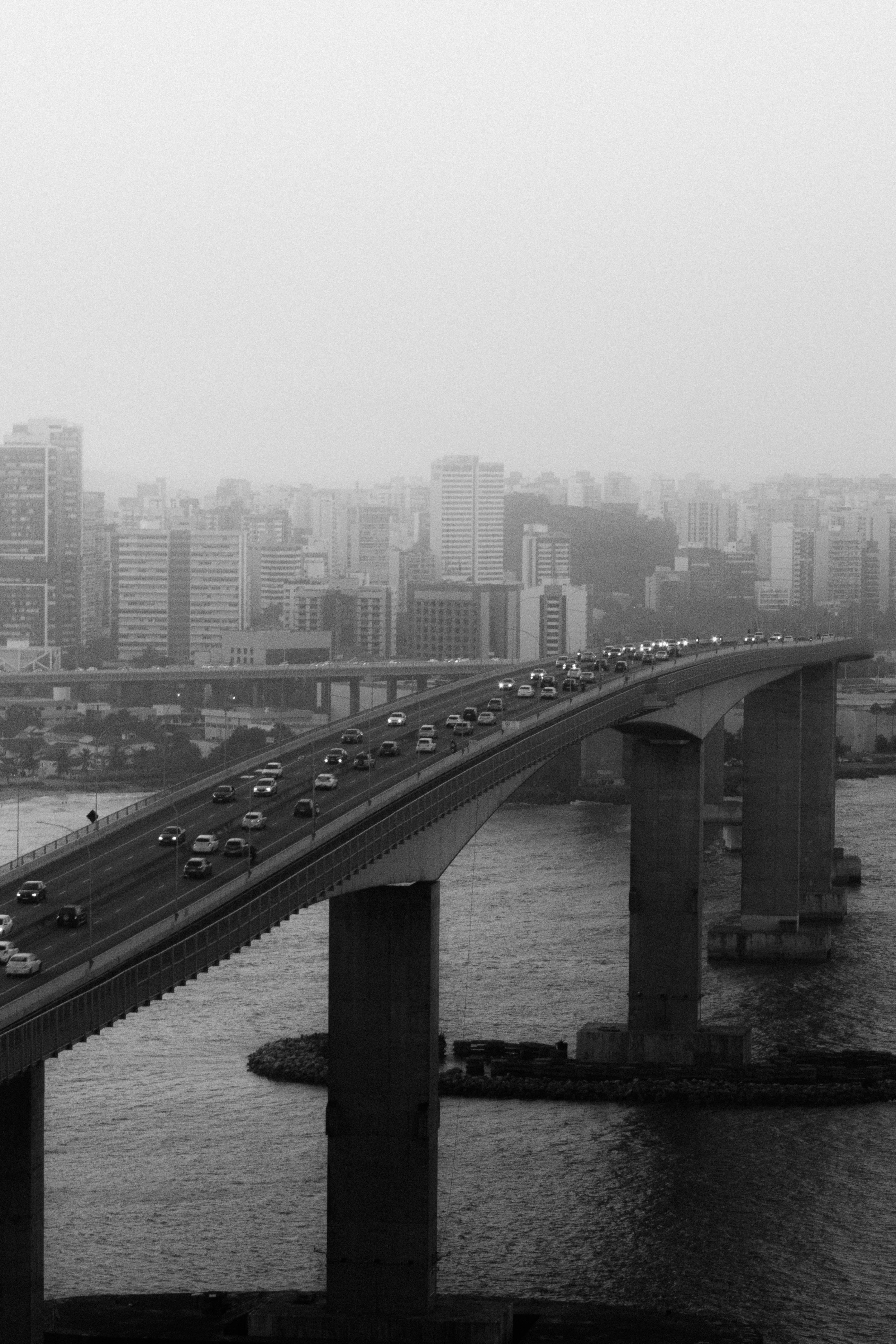 Black and white view of Vitoria, Brazil, showing a bridge over a foggy river amid cityscape.