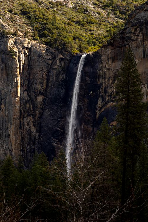 Free Stunning waterfall drops from a rocky cliff surrounded by lush trees in a serene outdoor setting. Stock Photo