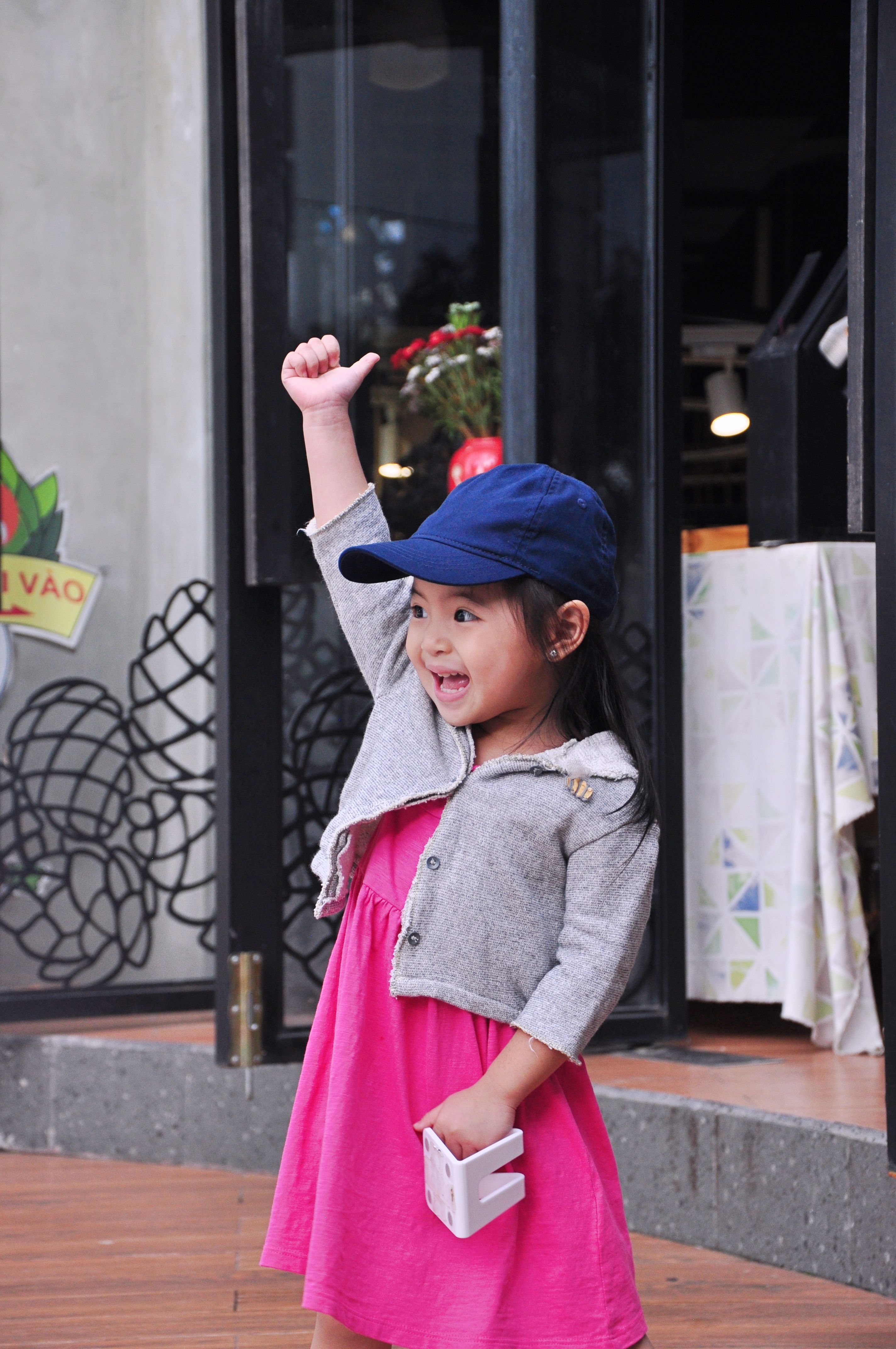 A cheerful young girl in a pink dress and blue cap smiling outdoors.